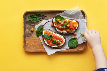 Breakfast starts a brisk day. Sandwich with slightly salted salmon, chickpeas and seasonal herbs. Healthy eating concept. On the tray is a snack, a yellow bright background, the child reaches for food