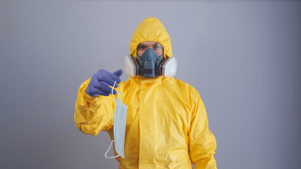 A man in yellow chemical protection suit and a respirator holds out a medical mask a respirator for protection against viruses, a gray background.