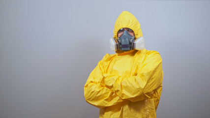 A man in yellow Hazmat suits and a respirator stands on a gray background with his arms crossed over his chest.