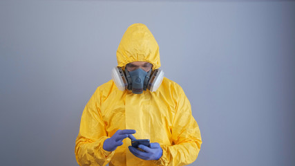 A man in yellow chemical protection suit and a respirator uses a phone gray background.