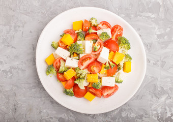 Vegetarian salad with broccoli, tomatoes, feta cheese, and pumpkin on white ceramic plate on a gray concrete background, top view.