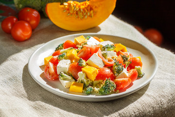 Vegetarian salad with broccoli, tomatoes, feta cheese, and pumpkin on white ceramic plate on linen textile, side view.