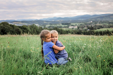 Naklejka na ściany i meble Cute 7-8 years girl hugging her smaller on the top of the mountains in summer