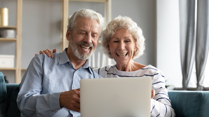 Overjoyed mature 60s husband and wife sit relax on sofa in living room have fun watch funny video on laptop, smiling elderly couple rest on couch browse internet use modern computer at home together