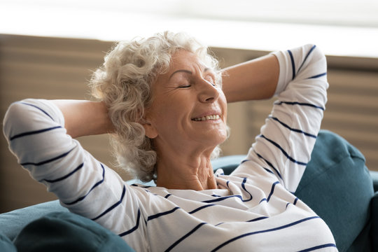 Smiling middle-aged 50s woman rest on comfortable sofa in living room with eyes closed take nap at home, happy elderly 60s wife relax on cozy couch sleep dream visualize, stress free concept