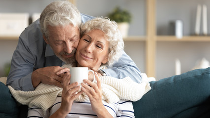Caring elderly 60s husband caress hug middle-aged wife bring tea cover with blanket at home, happy...