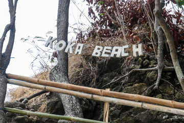 A sign 'Koka Beach' hanging inbetween the trees, at the entrance to the headland viewing point on Flores, Indonesia. There is a bit of overcast. Barren trees. The pathway is secured with bamboo rail.