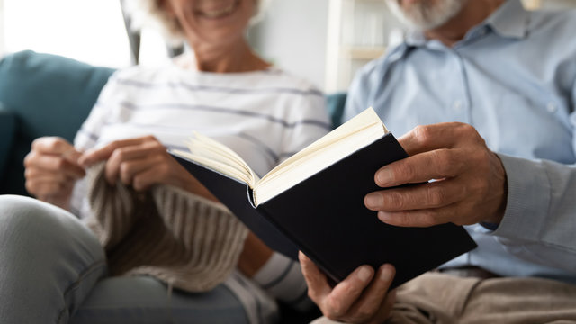 Close Up Of Happy Retired Elderly Couple Spouses Sit Relax On Couch Enjoy Leisure Weekend At Home Together, Smiling Mature 60s Husband Reading Book In Living Room, Middle-aged 50s Wife Do Knitting