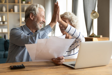 Happy old 60s couple spouses sit at desk at home give high five celebrate good news in letter document, smiling elderly husband and wife pay bills on internet on laptop online, easy banking system - obrazy, fototapety, plakaty
