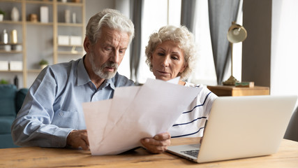 Focused elderly husband and wife sit at table in living room manage household document pay bills on...