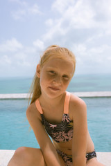A small and beautiful girl with red hair and freckles sits by the pool in a bathing suit in the villa and poses in front of the camera. Children's fashion