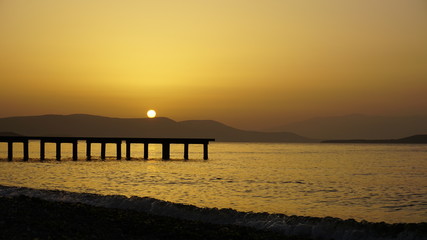 Fototapeta premium Dramatic sunset landscape at Urla, Izmir, Turkey. Beautiful blazing sunset over bright blue sea, pier, orange & purple sky above it with awesome golden rays of sun light reflection on calm waves. 
