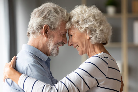Happy mature 50s couple embrace touch foreheads look in the eyes show love and care at home, smiling elderly 60s husband and wife hug cuddle share close tender romantic moment together