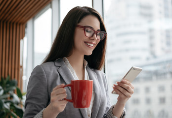 Businesswoman holding cellphone and cup of coffee, communication, checking email in office. Beautiful woman in stylish red eyeglasses using mobile app for online shopping. Freelancer receive payment