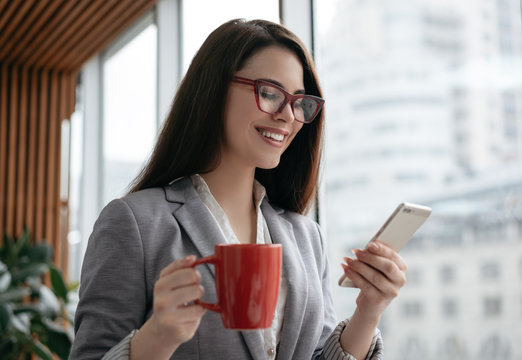 Businesswoman Holding Cellphone And Cup Of Coffee, Communication, Checking Email In Office. Beautiful Woman In Stylish Red Eyeglasses Using Mobile App For Online Shopping. Freelancer Receive Payment