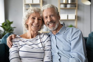 Headshot portrait of smiling elderly 60s husband and wife sit relax on couch hugging cuddling, happy mature old couple rest on sofa in living room embrace look at camera show love and care