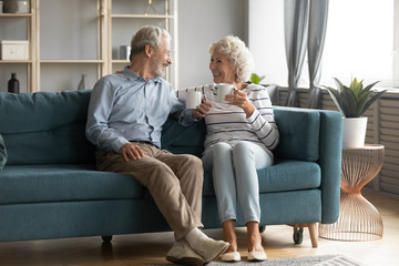 Happy mature 50s husband and wife sit rest on comfortable sofa in living room enjoy tea talking, smiling elderly 60s couple relax on couch at home drink coffee chat speak laugh on leisure weekend