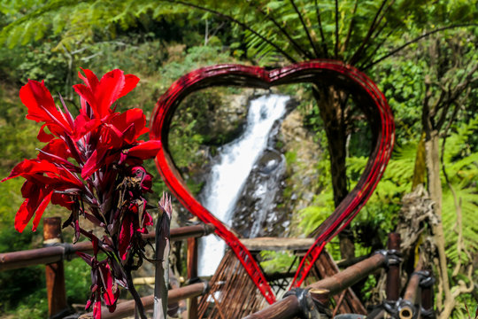 A red heart-shaped frame with Gitgit Twin Waterfall in the back, Bali Indonesia. There are red flowers. Touristic attraction for taking selfies with the waterfall. Lush green flora of the jungle.