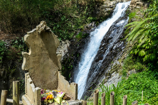 An offering altar with on Gitgit Twin Waterfall, Bali, Indonesia. The waterfall is surrounded by lush green flora, in a dense jungle. The altar resembles a throne, few offerings on it.