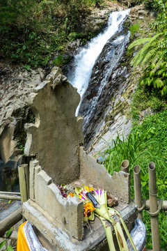 An offering altar with on Gitgit Twin Waterfall, Bali, Indonesia. The waterfall is surrounded by lush green flora, in a dense jungle. The altar resembles a throne, few offerings on it.