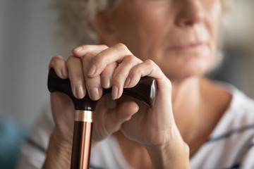 Close up of pensive mature disabled woman with walking stick lost in thoughts feeling abandoned lonely at home, sad old 60s grandmother with cane thinking mourning indoors, elderly solitude concept