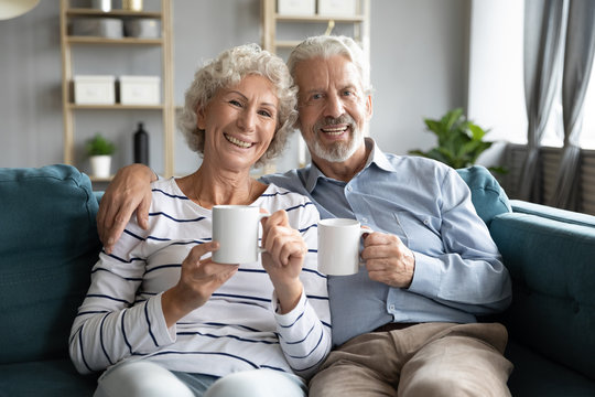 Family Portrait Of Elderly 60s Husband And Wife Sit Relax On Couch In Living Room Hugging Cuddling Look At Camera, Happy Mature 50s Couple Rest On Sofa Drink Hot Tea Enjoy Weekend At Home Together