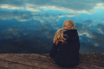 young girl with long blonde hair is sitting on a wooden stage while looking over the rippled water with reflecting cloudy sky in a thoughtful pose - coming of age, outdoor and nature care concept - Powered by Adobe