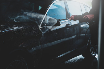 Hands of a male washing black car outdoors in a carwash station, using water jet with soap and high...