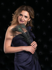 beautiful smiling woman with long hair and makeup in purple bare shoulder dress with bow holding peacock feathers and looking at camera on black