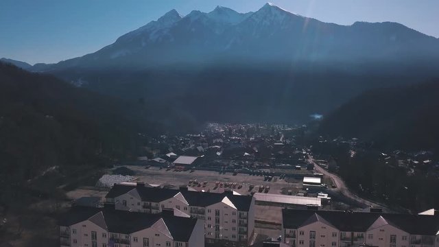 Aerial view of the small town situated in the valley surrounded by high mountains covered by trees. Clip. Flying above the town near forested hills on blue sky background.