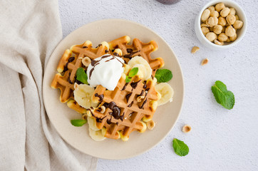 Viennese waffles with fresh banana, hazelnuts, chocolate sauce and whipped cream on a plate on a light background. Delicious breakfast. Horizontal orientation. Close up. Top view. Selective focus.