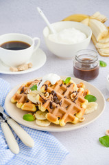 Viennese waffles with fresh banana, hazelnuts, chocolate sauce and whipped cream on a plate on a light background. Delicious breakfast. Vertical orientation. Selective focus.