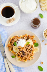 Viennese waffles with fresh banana, hazelnuts, chocolate sauce and whipped cream on a plate on a light background. Delicious breakfast. Vertical orientation. Selective focus. Top view.