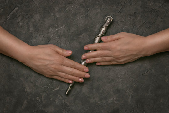 Beautiful female hands with pink manicure hold a tool: a large-diameter drill, on a background with gray paint. The contrast is male and female, light and dark. Industrial still life.