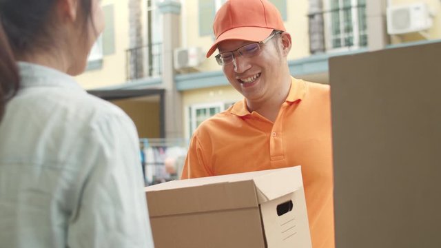 Young postal delivery courier man holding parcel boxes for sending to customer, Asian women signing and pay via qr code on mobile phone for receive delivered parcels outdoors. 4k Slow motion.