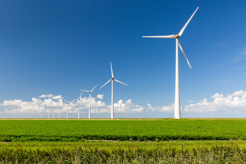 A long row of wind turbines emerging from green grass and against a summer blue sky with distant clouds