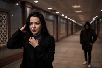 a young beautiful woman walks through an underground passage at night, followed by a man in dark...
