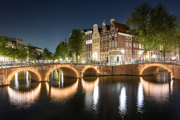 Fototapeta na wymiar Wide angle cityscape of Amsterdam at night, with an historical bridge lit with nightlights reflecting on waters
