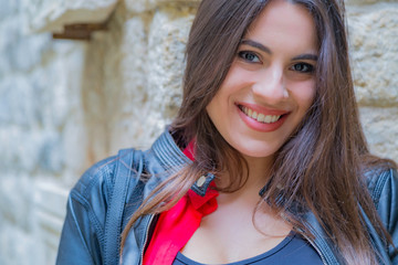 Bellissima donna italiana con un sorriso solare e capelli lunghi castani lisci indossa una camicia rossa e giacca nera di pelle in esterno appoggiata ad un muro di pietra di una città medievale