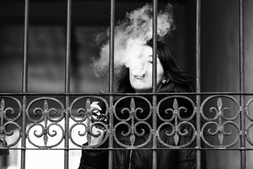 Vape teenager. Young pretty caucasian brunette girl smoking an electronic cigarette near old metal gate on the street in the spring. Deadly bad habit. Black and white.