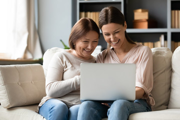 Happy older mature mother sitting on sofa with excited grownup daughter, looking at laptop screen. Laughing young woman showing funny video photos to elderly mommy, having fun together at home.