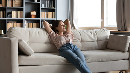 Calm happy sleepy woman crossed hands behind head, relaxing on comfortable sofa alone in living...