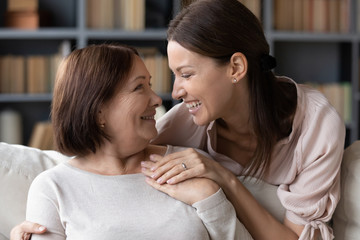 Obraz na płótnie Canvas Laughing young woman embracing from back sitting on sofa smiling older mother. Affectionate grownup daughter sharing news, communicating talking to pleasant middle aged mature mommy at home.