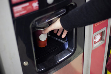 Hot coffee from the vending machine. A hand reaches for a hot cup with a drink obtained from an automatic device. Coffee at the mall. A girl takes a drink from a special compartment in the device.