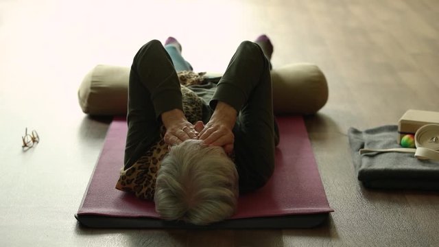 Group of woman in yoga session lying in corpse pose, with knee support of bolster covering eyes with hand while relaxing and resting after workout
