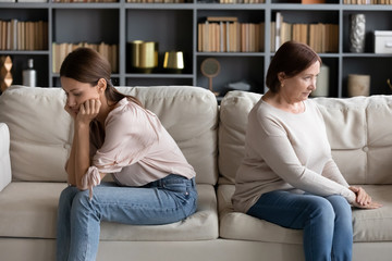 Stressed young woman sitting separately with offended mature old mother in living room. Unhappy...