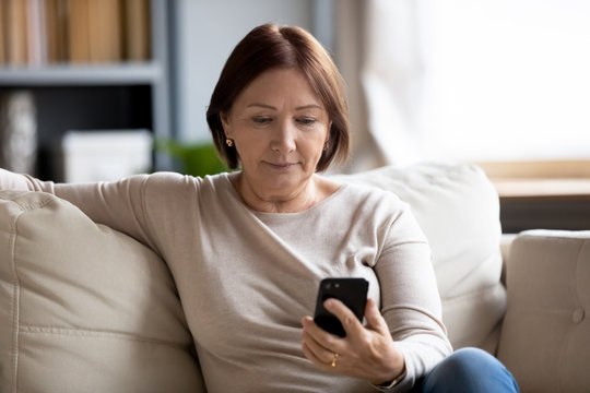 Worried Middle Aged Woman Sitting On Couch, Looking At Mobile Phone Screen. Serious Senior Mature Mother Waiting For Response From Grownup Children, Chatting In Social Network Online Via Smartphone.