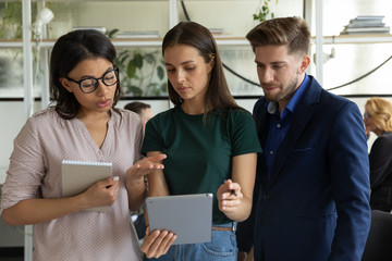 Concentrated multiracial businesspeople stand brainstorm using modern tablet gadget, discuss ideas at workplace, focused divers multiethnic colleagues talk cooperate in office, collaboration concept