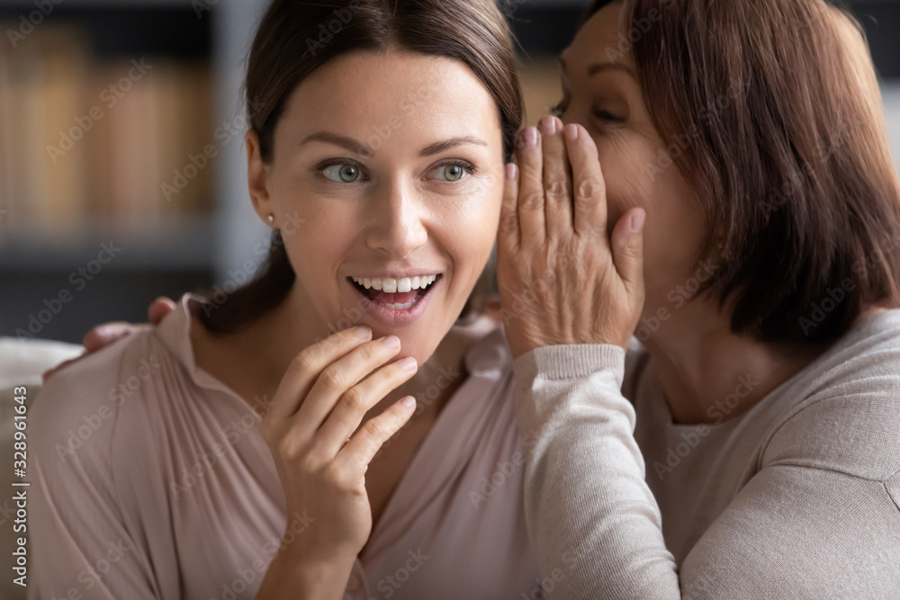 Wall mural Head shot curious young woman listening to secret information from senior older mommy. Happy middle aged mother whispering gossip, spreading rumor to grownup daughter, having fun together at home.