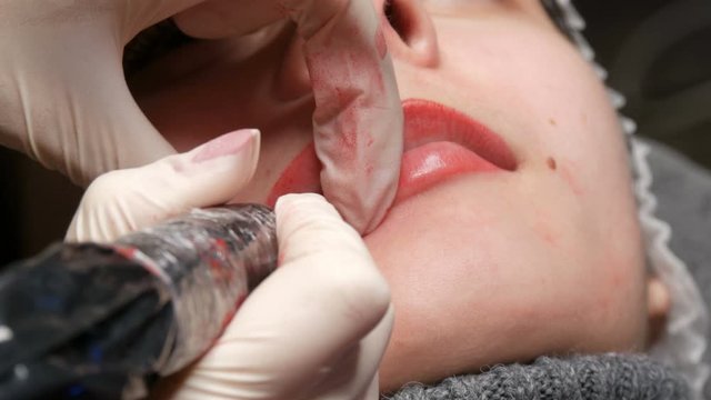 Microblading lip tattoo with a special coloring red pigment that corrects lip color in a cosmetology clinic. Permanent makeup lips procedure applying pigment makeup on lips with a tattoo machine close